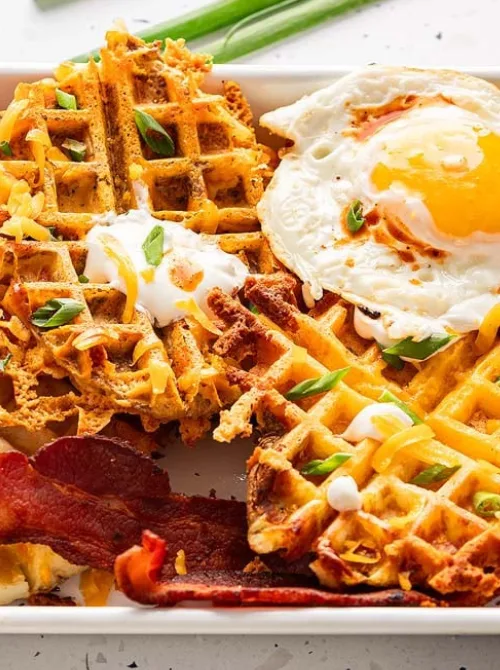 Plate of crispy savory waffles made from leftover ingredients, topped with a fried egg, shredded cheese, sour cream, and green onions—an inventive and delicious twist for creative leftovers recipes.