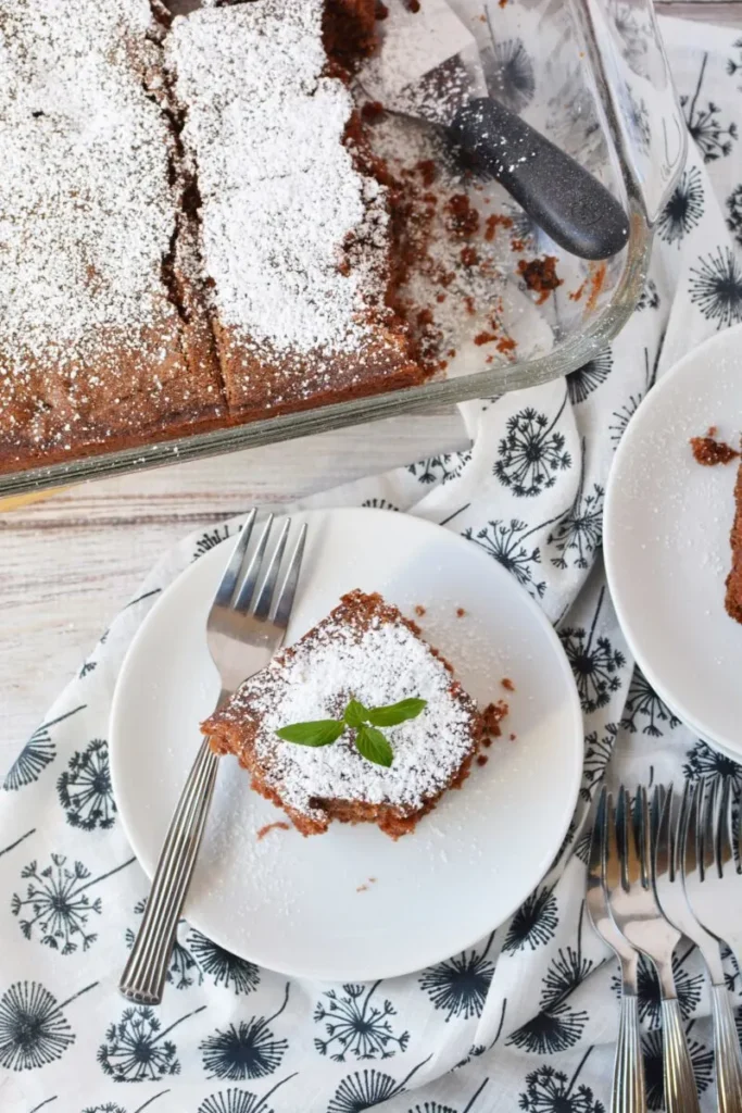 Square slice of chocolate cake dusted with powdered sugar and garnished with a fresh mint leaf, served on a white plate—a delightful dessert that can be made using leftovers recipes.