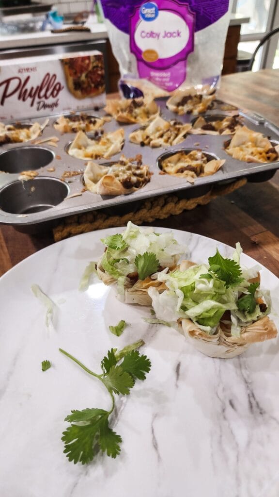 Mini taco cups made with crispy phyllo dough, filled with seasoned ground meat, beans, and melted cheese, topped with shredded lettuce and fresh cilantro. Displayed on a plate and in a muffin tin, these bite-sized treats are a fun and flavorful Superbowl appetizer.