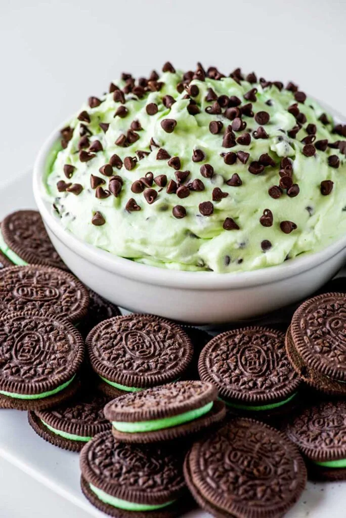 A bowl of creamy mint chocolate chip dip topped with mini chocolate chips, surrounded by mint Oreo cookies, perfect for St Patrick's Day desserts or festive Irish treats.