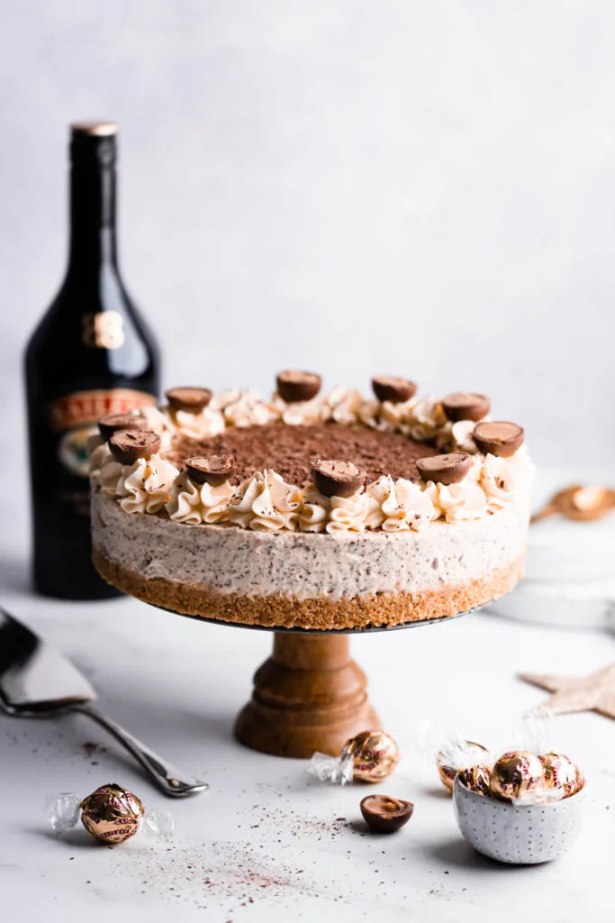 A creamy Bailey's cheesecake topped with whipped cream swirls, chocolate truffles, and grated chocolate, displayed on a wooden stand, perfect for Irish desserts or St Patrick's Day celebrations.