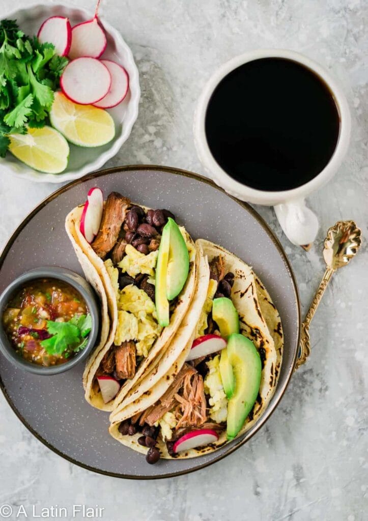 Breakfast tacos filled with scrambled eggs, black beans, avocado slices, and pulled pork leftovers, served with fresh lime wedges, radishes, salsa, and a cup of black coffee.