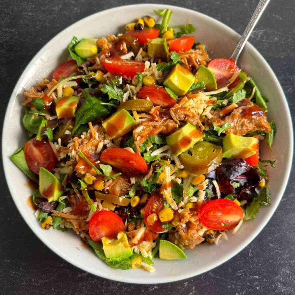 Colorful salad topped with pulled pork leftovers, cherry tomatoes, avocado chunks, shredded cheese, corn, jalapeños, and a drizzle of barbecue sauce, served in a white bowl.