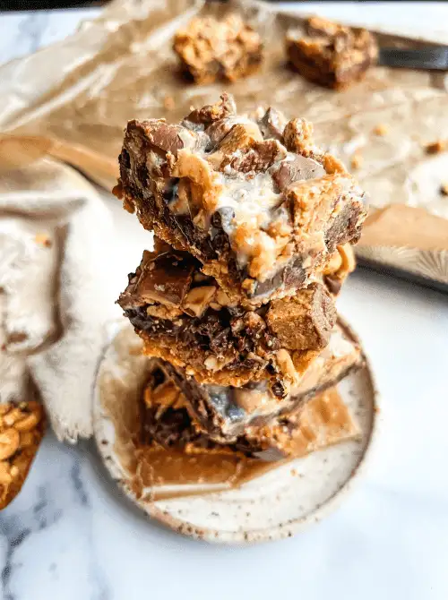 Stack of gooey layered dessert bars loaded with chopped chocolate, nuts, and drizzled sweetened condensed milk on a small ceramic plate—perfect for indulgent snacking.