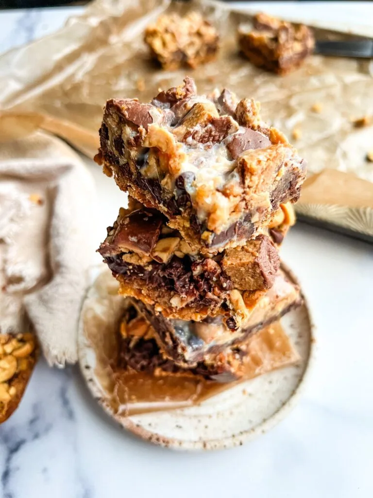 Stack of gooey chocolate peanut butter bars filled with caramel and chunks of candy, served on a small plate. A rich and indulgent Super Bowl food idea for game day desserts.