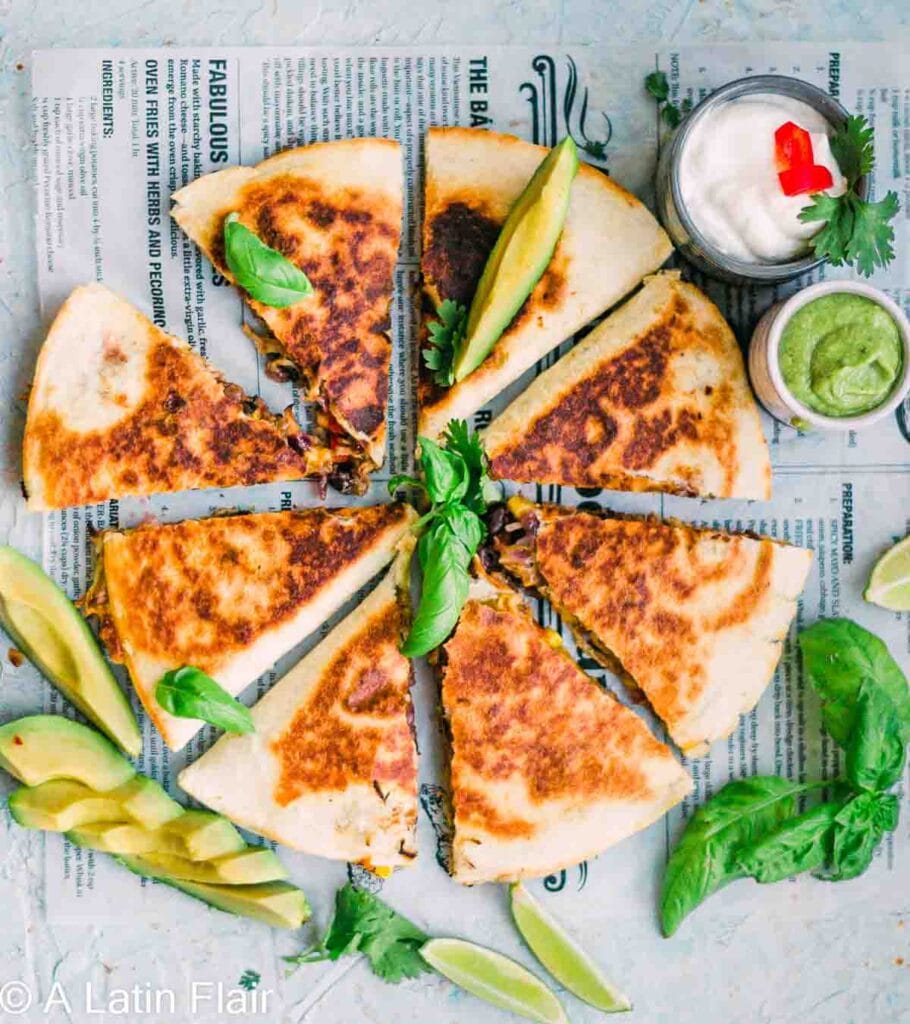 Golden, crispy quesadilla slices arranged in a circle, garnished with fresh avocado, basil, and lime wedges, served with sides of sour cream and green salsa—a flavorful and creative way to repurpose leftovers.