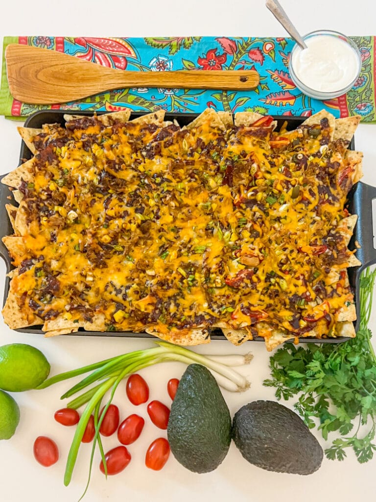 Sheet pan nachos loaded with leftover pulled pork, melted cheddar cheese, and green onions, surrounded by fresh ingredients like avocados, limes, cherry tomatoes, and cilantro, with a side of sour cream.