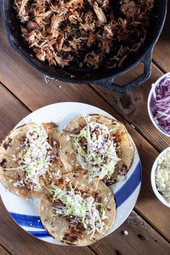 Plate of shredded pork tacos topped with fresh cabbage, red onions, and crumbled cheese, served alongside a skillet of pulled pork—an easy and delicious choice for leftovers recipes.