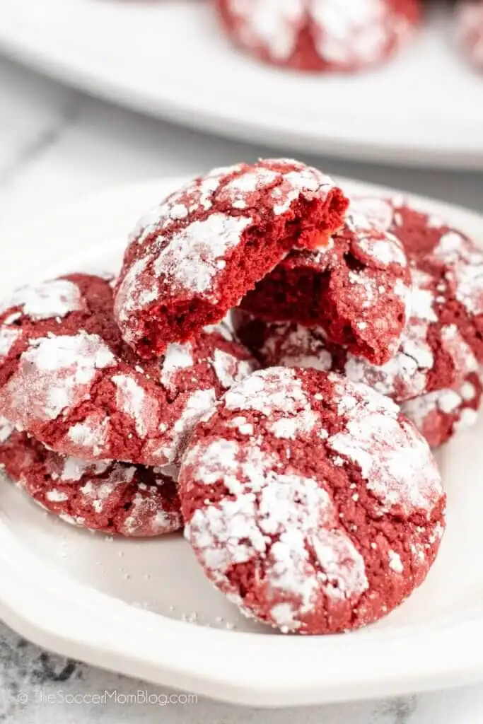 A plate of vibrant red heart cookies coated in a dusting of powdered sugar, with one cookie broken in half to reveal a soft, chewy center.