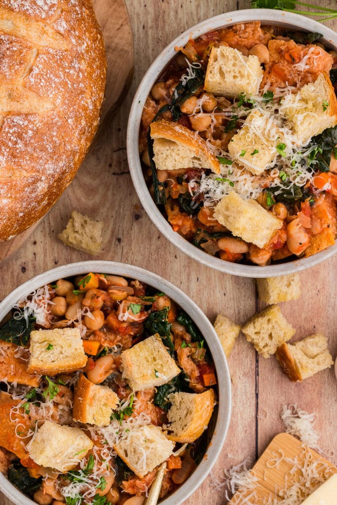 Bowls of hearty vegetable and bean stew topped with crispy bread cubes and shredded cheese, served alongside a loaf of crusty bread—a warm and satisfying option for leftovers recipes.