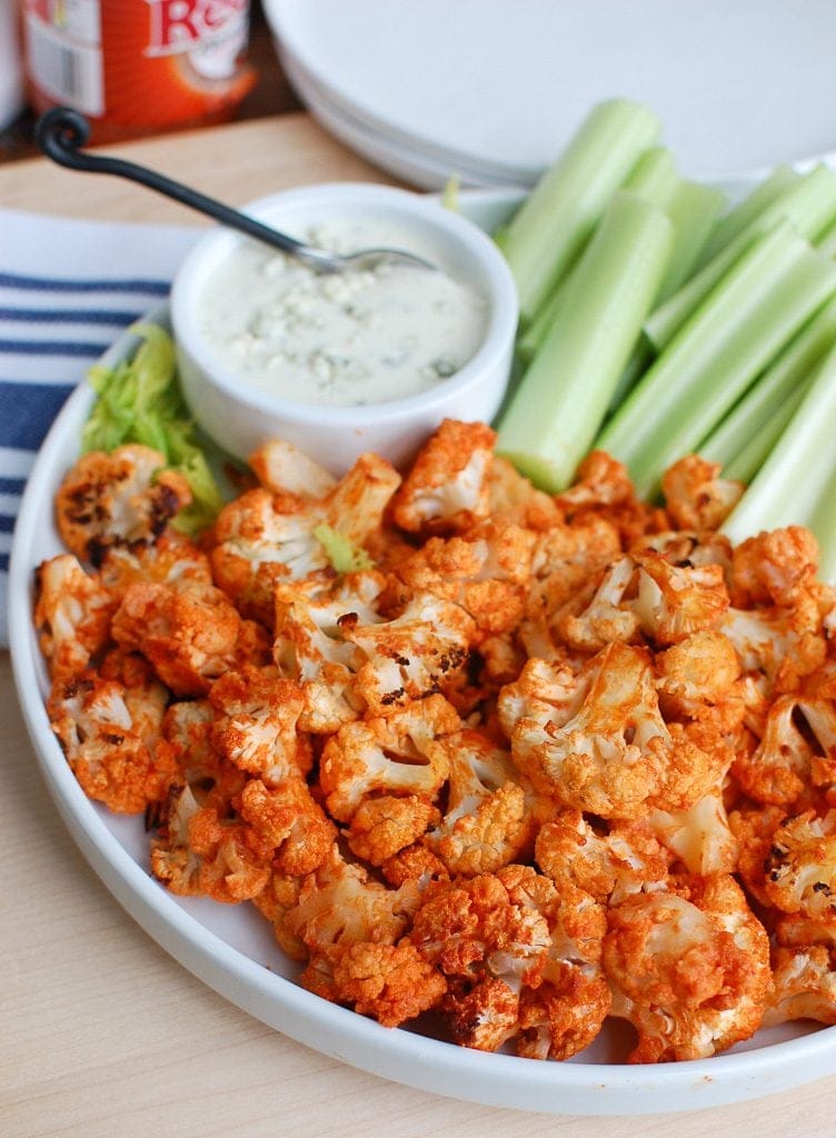 A plate of roasted Buffalo cauliflower bites coated in spicy Buffalo sauce, served with fresh celery sticks and a bowl of creamy blue cheese dressing for dipping. This healthy and flavorful dish is a perfect Superbowl appetizer for vegetarians and spice lovers alike.
