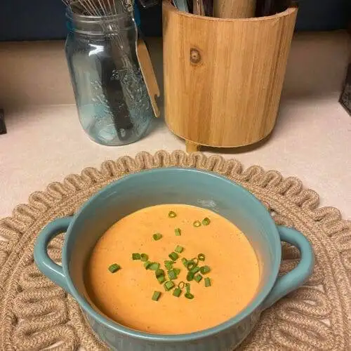 A bowl of creamy Roasted Red Pepper & Sesame Soy Dip garnished with fresh chopped chives, served in a blue dish on a textured placemat, with kitchen utensils in the background.
