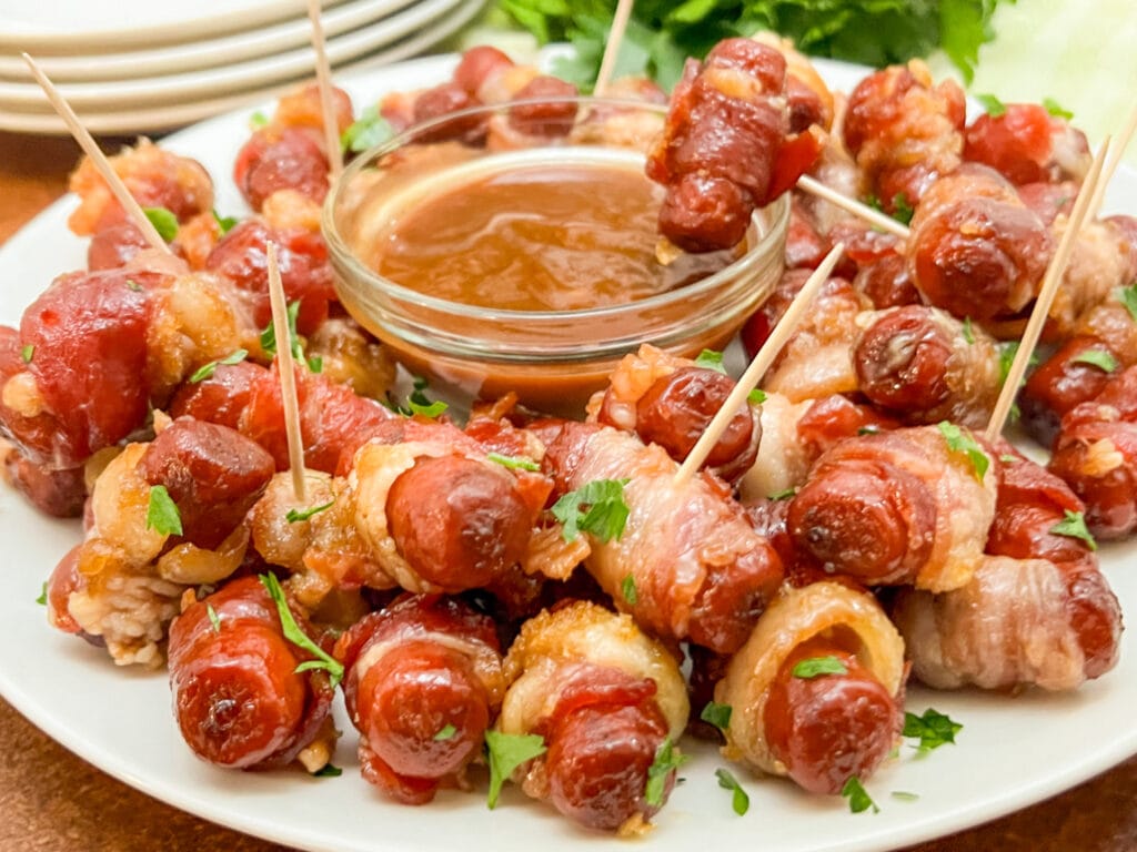 A plate of bacon-wrapped smokies, glazed with a sweet and savory sauce and garnished with chopped parsley. Served with a small bowl of dipping sauce, these bite-sized treats are a delicious and indulgent Superbowl appetizer.