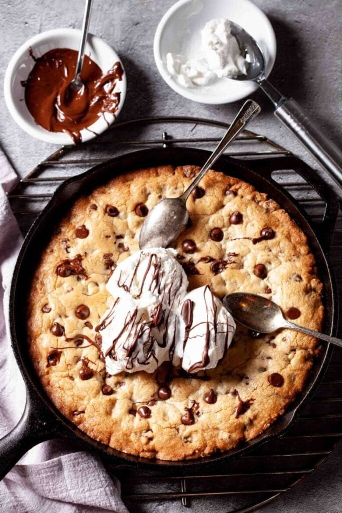 Skillet cookie loaded with chocolate chips, topped with scoops of vanilla ice cream drizzled with melted chocolate, surrounded by bowls of whipped cream and chocolate sauce, creating a fun and shareable Valentine's Cake Idea.