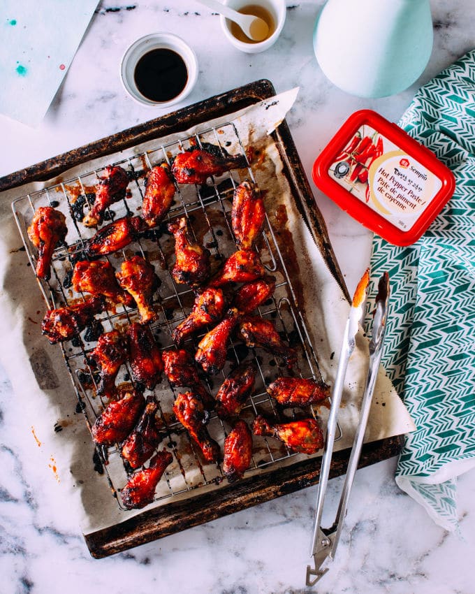 A tray of crispy baked Korean chicken wings resting on a wire rack over a baking sheet, brushed with a rich red glaze and slightly charred for a smoky flavor. The setup includes a small dish of soy sauce, a jar of Korean chili paste (gochujang), and kitchen tongs, set on a marble countertop with a teal patterned cloth. Perfect for Superbowl appetizers to spice up your game day spread.