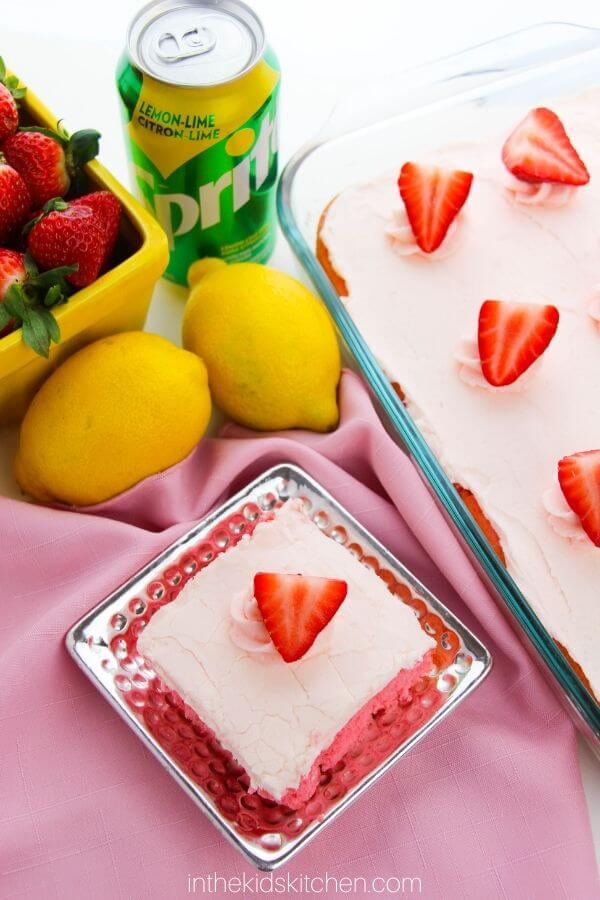 Bright and colorful setup featuring a lemon-strawberry sheet cake topped with pink frosting and fresh strawberry slices, accompanied by lemons, strawberries, and a can of Sprite. This dessert is a refreshing and creative option for Valentine's Cake Ideas.