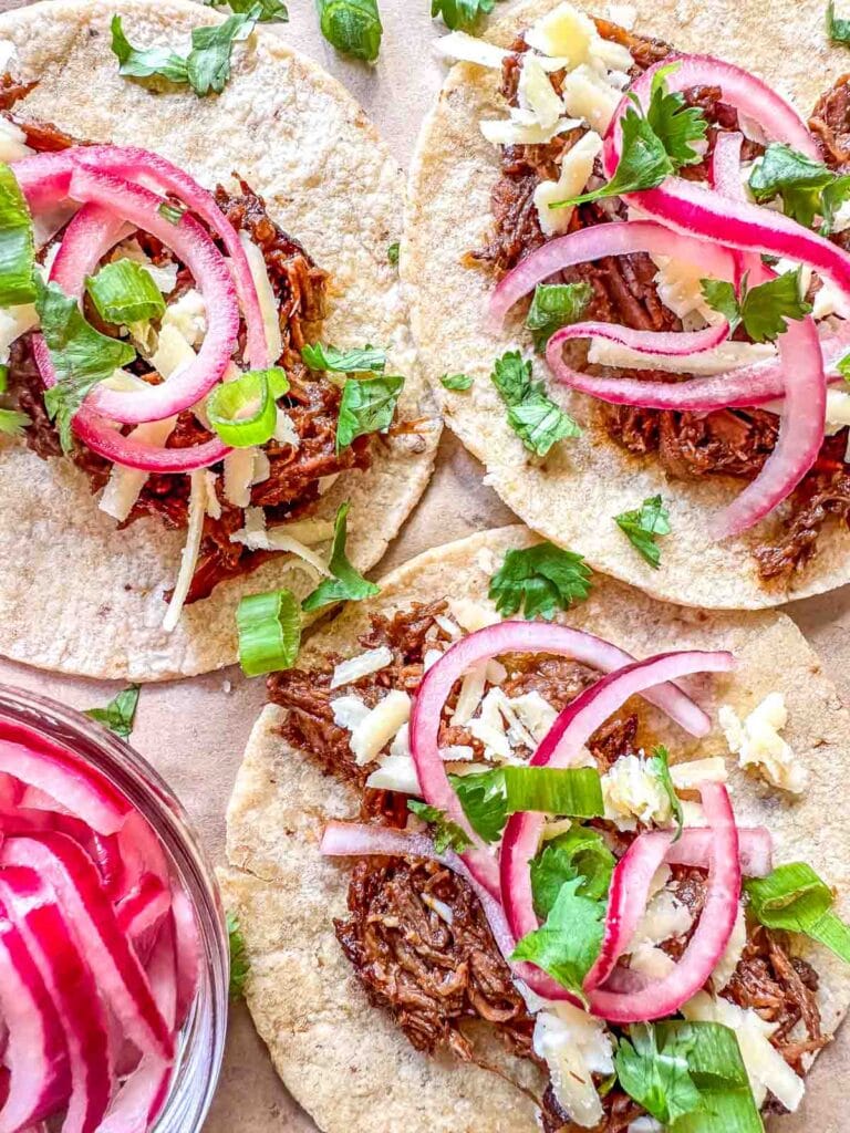 Soft corn tortillas topped with flavorful leftover pulled pork, pickled red onions, shredded cheese, and fresh cilantro, served on a parchment-lined surface.