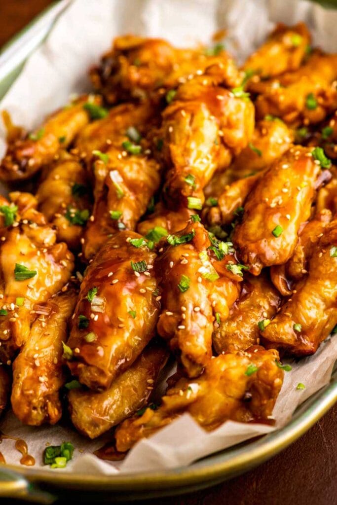 A platter of sticky chicken wings glazed with a flavorful sauce and garnished with chopped green onions and sesame seeds. Served in a lined dish, these wings are a mouthwatering option for a Superbowl appetizer or party snack.