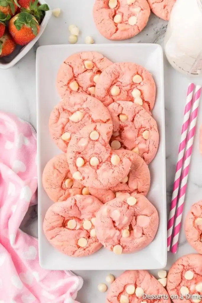 A white rectangular plate filled with soft pink heart cookies, studded with creamy white chocolate chips, creating a charming and festive dessert. The plate is surrounded by a pink polka-dot cloth, fresh strawberries, and striped straws, perfect for a sweet Valentine's Day treat.