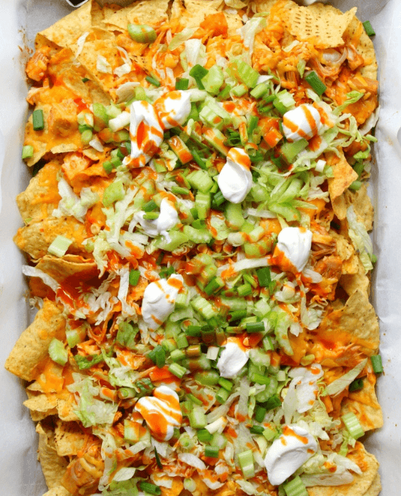 A tray of Buffalo chicken nachos loaded with shredded chicken tossed in Buffalo sauce, melted cheese, shredded lettuce, diced celery, green onions, and dollops of sour cream. Drizzled with additional Buffalo sauce for extra flavor, this zesty dish makes for a crowd-pleasing Superbowl appetizer.