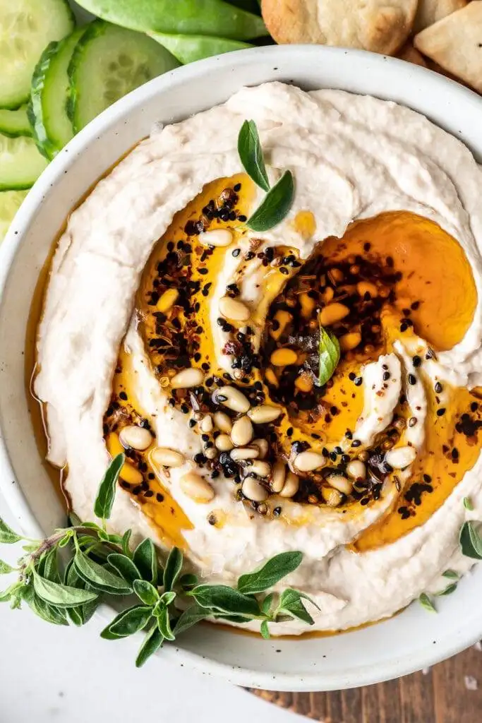 Luxurious hummus drizzled with olive oil, sprinkled with pine nuts, herbs, and spices, served with fresh cucumber slices and pita bread—a standout choice among the best party dips.