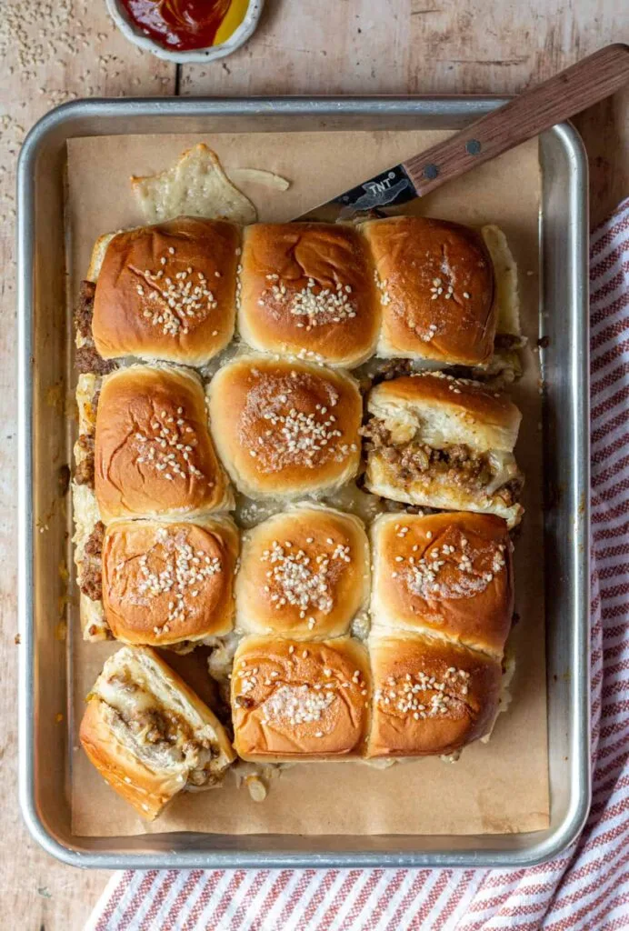 A tray of freshly baked cheeseburger sliders topped with sesame seeds, served on parchment paper. A savory and budget-friendly Super Bowl food idea for game day feasts.