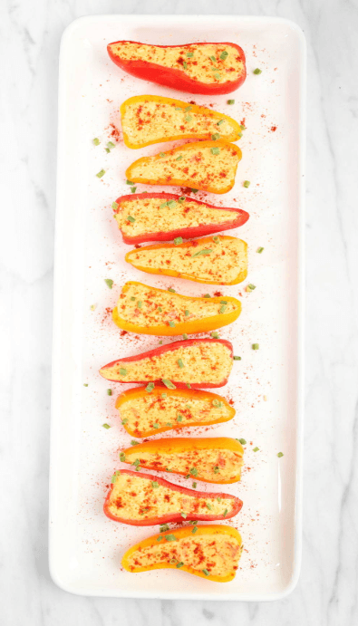A platter of colorful mini bell peppers halved and stuffed with a creamy filling, sprinkled with paprika and garnished with chopped chives. These vibrant and bite-sized snacks are an elegant and healthy Superbowl appetizer.