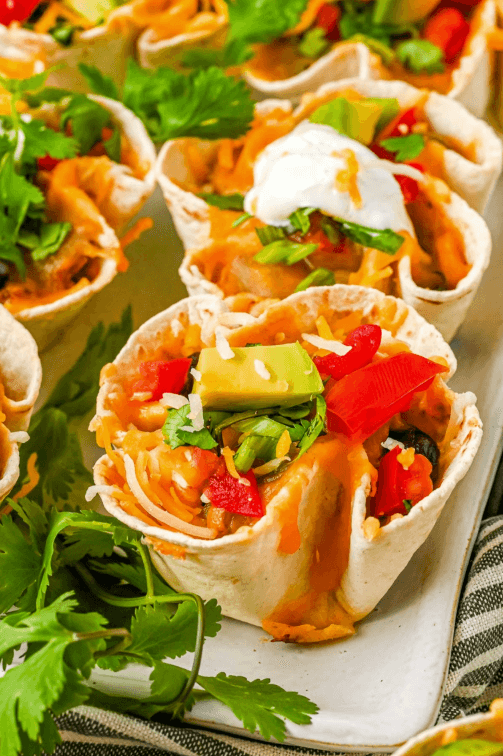 Mini taco cups made with soft tortillas and filled with layers of melted cheese, diced tomatoes, avocado, sour cream, and fresh cilantro. These colorful and bite-sized snacks are perfect for a festive Superbowl appetizer.