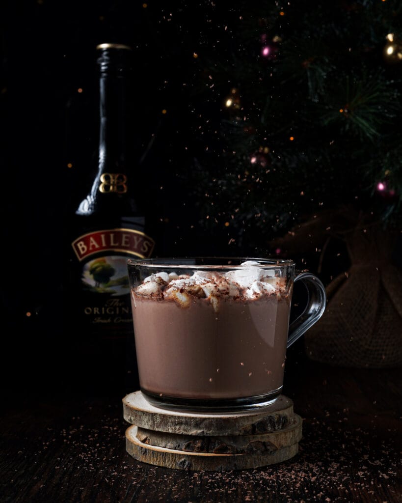 A glass mug of rich hot chocolate topped with marshmallows and cocoa powder, set on rustic wooden coasters, with a bottle of Baileys in the background and festive lights, creating a cozy atmosphere for boozy treats.