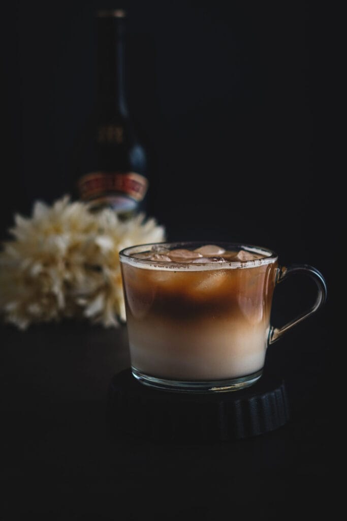 A layered iced coffee cocktail served in a glass mug, with creamy milk at the bottom and rich coffee on top, set against a dark background with blurred flowers and a bottle of Baileys, perfect for indulgent boozy treats.