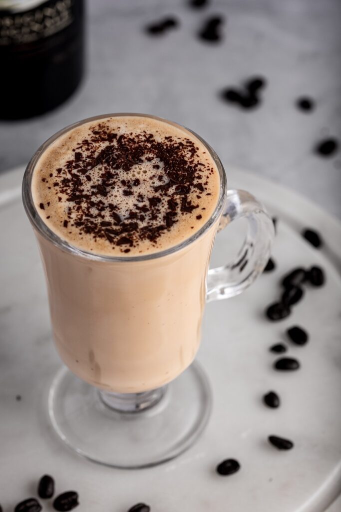 A glass mug filled with a creamy coffee cocktail topped with chocolate shavings, surrounded by scattered coffee beans on a white marble surface, perfect for indulgent boozy treats.