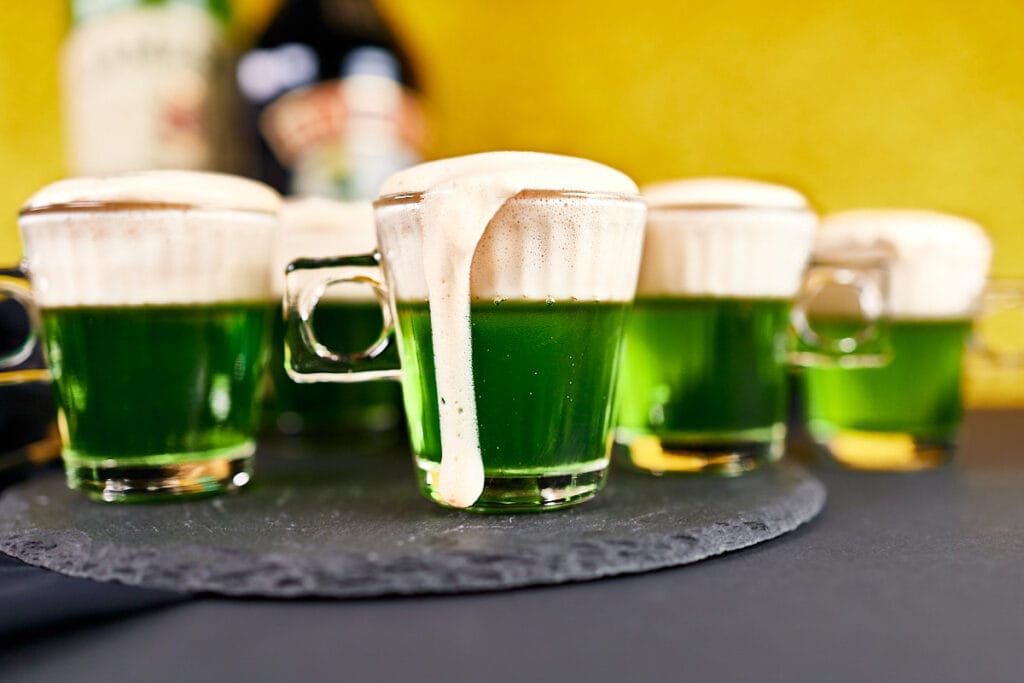 Small glass mugs filled with vibrant green drinks topped with frothy foam, some dripping down the sides, set on a slate serving tray with a blurred yellow and Baileys bottle in the background.