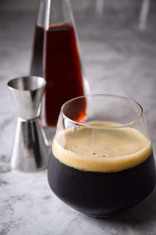 A glass of dark stout beer with a creamy foam head, accompanied by a silver jigger and a bottle of rich amber liquid in the background, set on a marble surface.