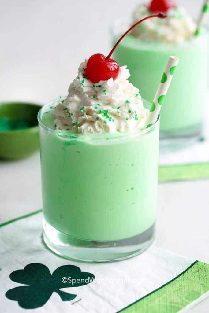 A festive green milkshake topped with whipped cream, green sprinkles, and a maraschino cherry, served with a green polka dot straw in a clear glass, placed on a napkin featuring a shamrock design.