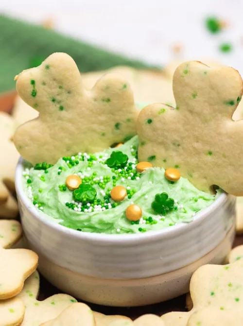 Shamrock-shaped cookies served with a bowl of green frosting dip decorated with festive sprinkles and gold accents, perfect for St. Patrick's Day desserts or Irish-themed treats.