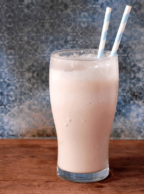 A tall glass filled with a creamy milkshake-like drink, featuring two blue and white striped straws, set against a rustic wooden surface and a textured blue background.