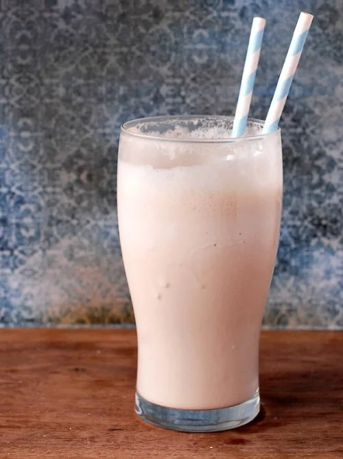 A tall glass filled with a creamy milkshake-like drink, featuring two blue and white striped straws, set against a rustic wooden surface and a textured blue background.