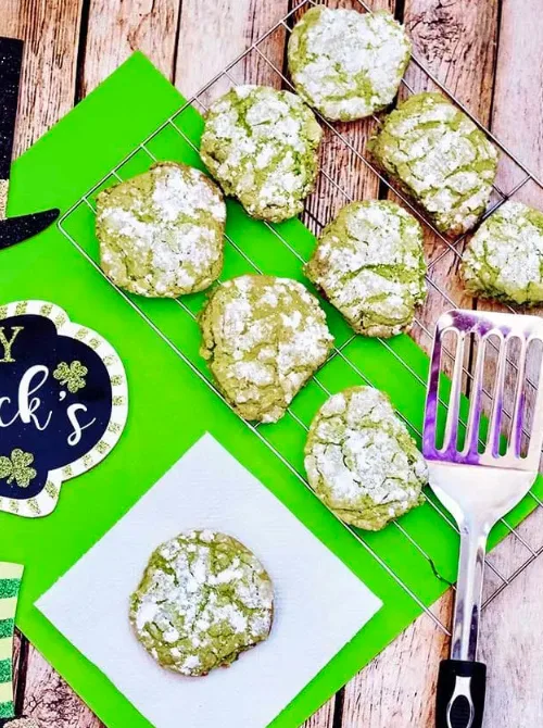 Green crinkle cookies dusted with powdered sugar cooling on a wire rack, surrounded by festive St. Patrick's Day decorations, perfect for Irish-inspired desserts.