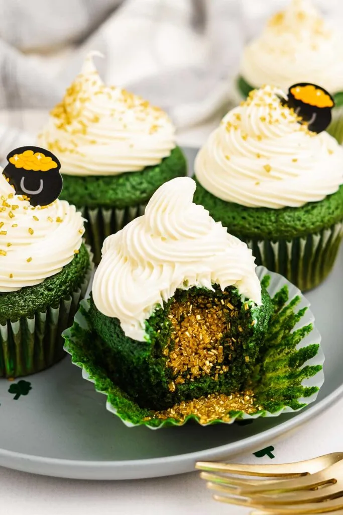 Green St. Patrick's Day cupcakes topped with creamy white frosting, gold sprinkles, and a pot of gold decoration, with one cupcake revealing a surprise golden sprinkle-filled center.