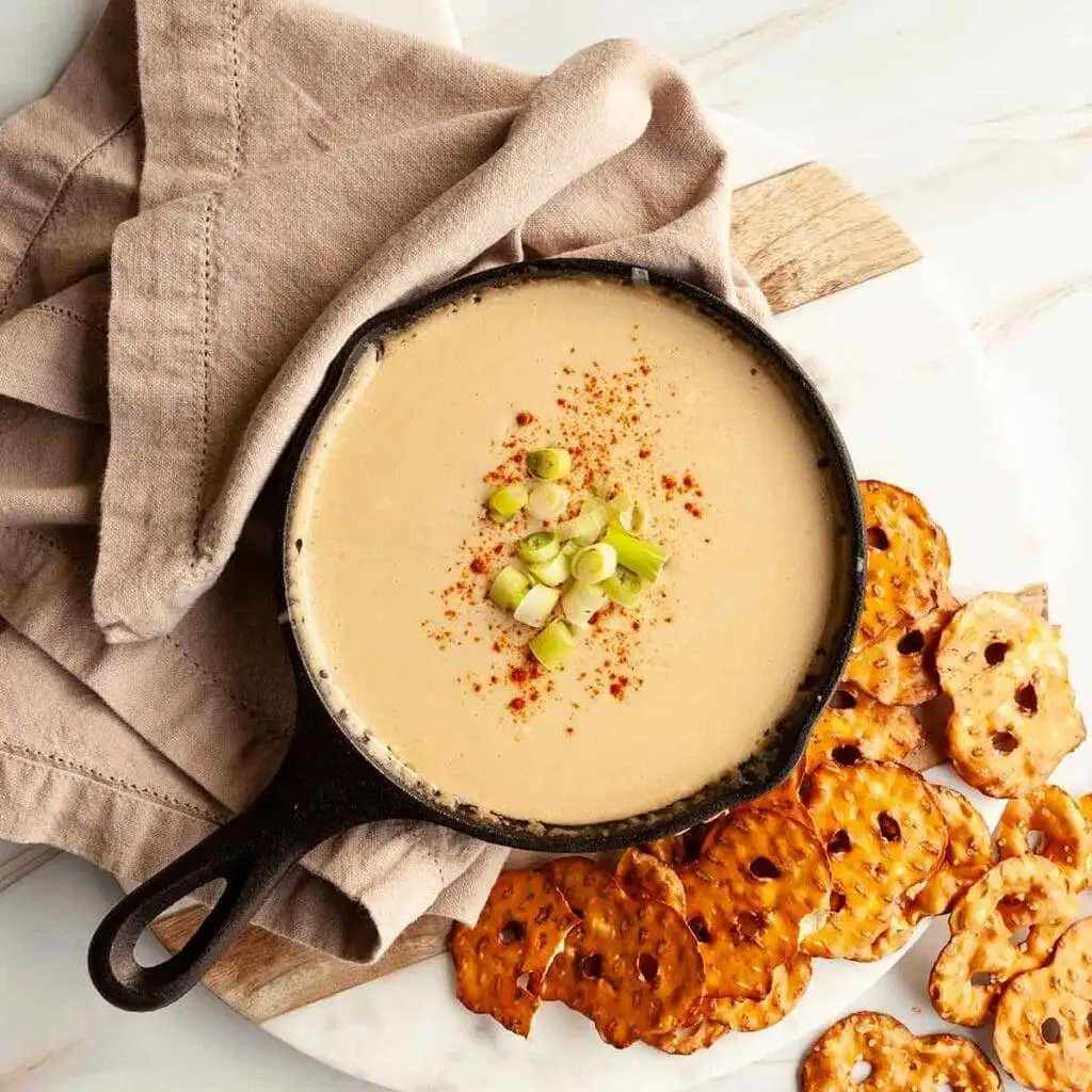 A skillet filled with creamy beer cheese dip, garnished with green onions and a sprinkle of paprika, served alongside crispy pretzel chips—perfect for Irish party food ideas.