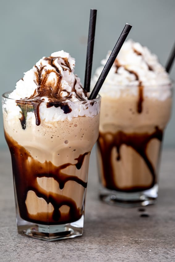 Two tall glasses filled with a creamy iced coffee drink, drizzled with chocolate syrup and topped with whipped cream, served with black straws, creating a decadent and indulgent treat.