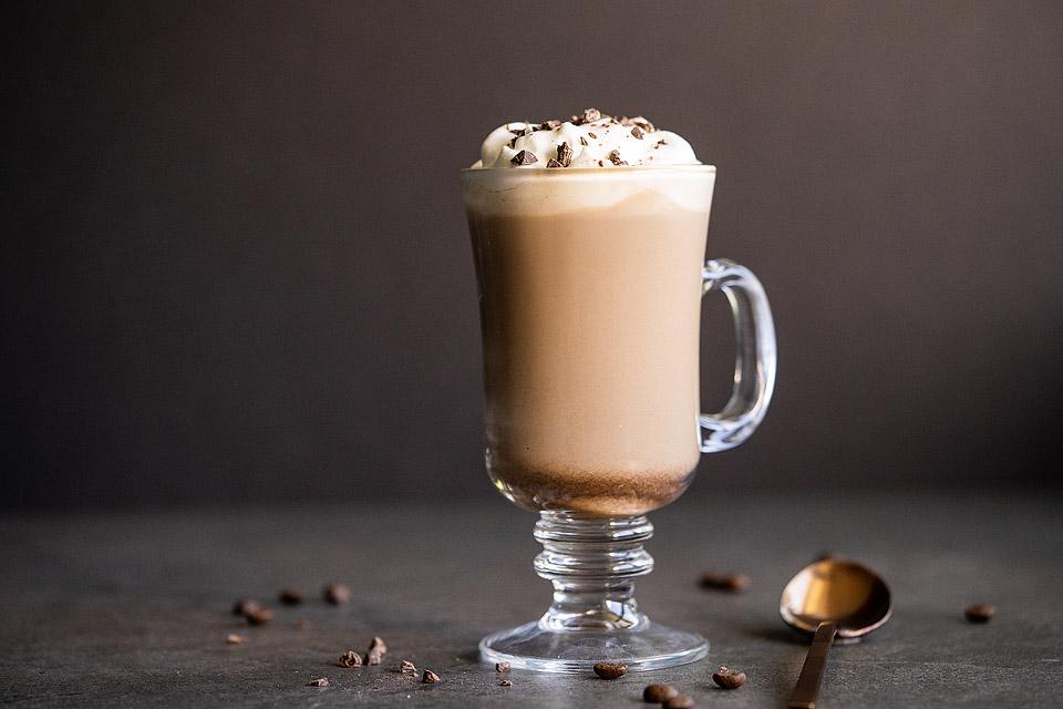 A glass mug filled with a creamy coffee cocktail, topped with whipped cream and chocolate shavings, surrounded by scattered coffee beans on a dark surface, perfect for enjoying as a boozy treat.