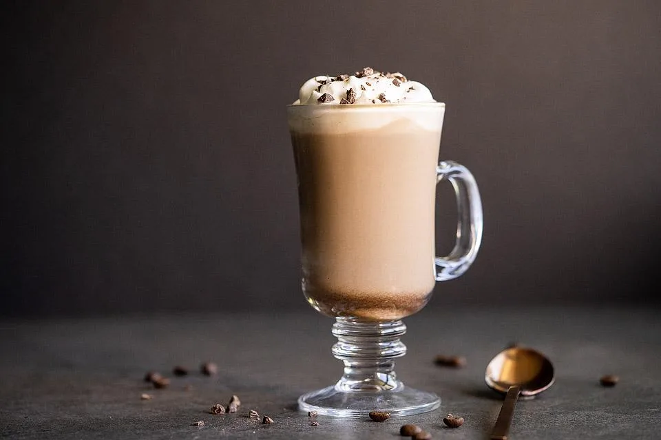 A glass mug filled with a creamy coffee cocktail, topped with whipped cream and chocolate shavings, surrounded by scattered coffee beans on a dark surface, perfect for enjoying as a boozy treat.