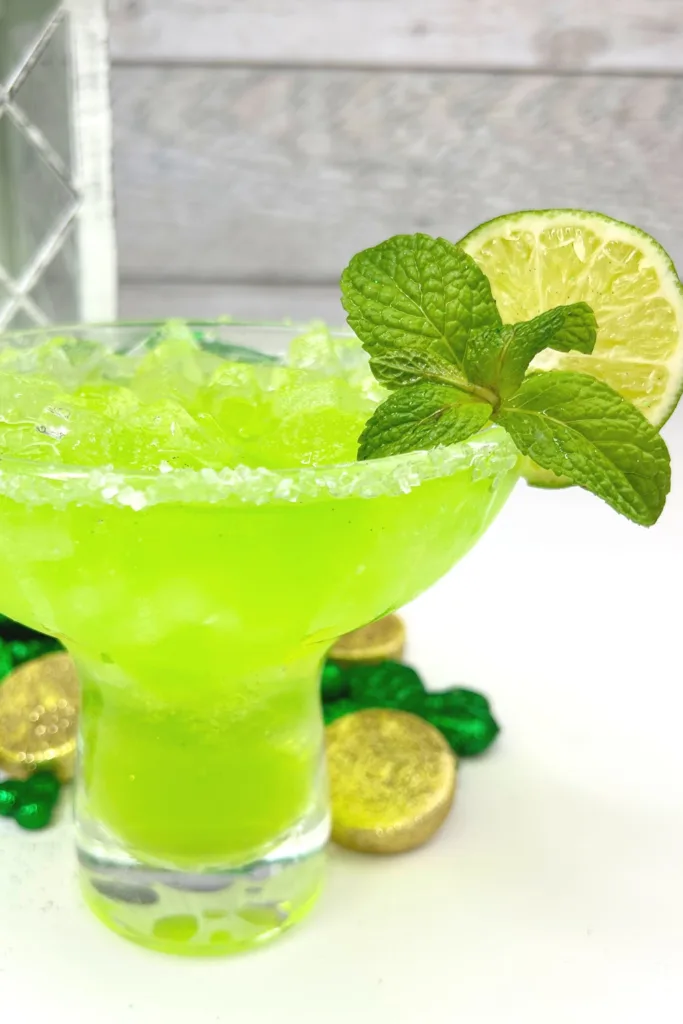 A vibrant green margarita in a salted-rim glass, garnished with a lime slice and fresh mint leaves, surrounded by festive gold coins and shamrocks, set on a bright white surface.