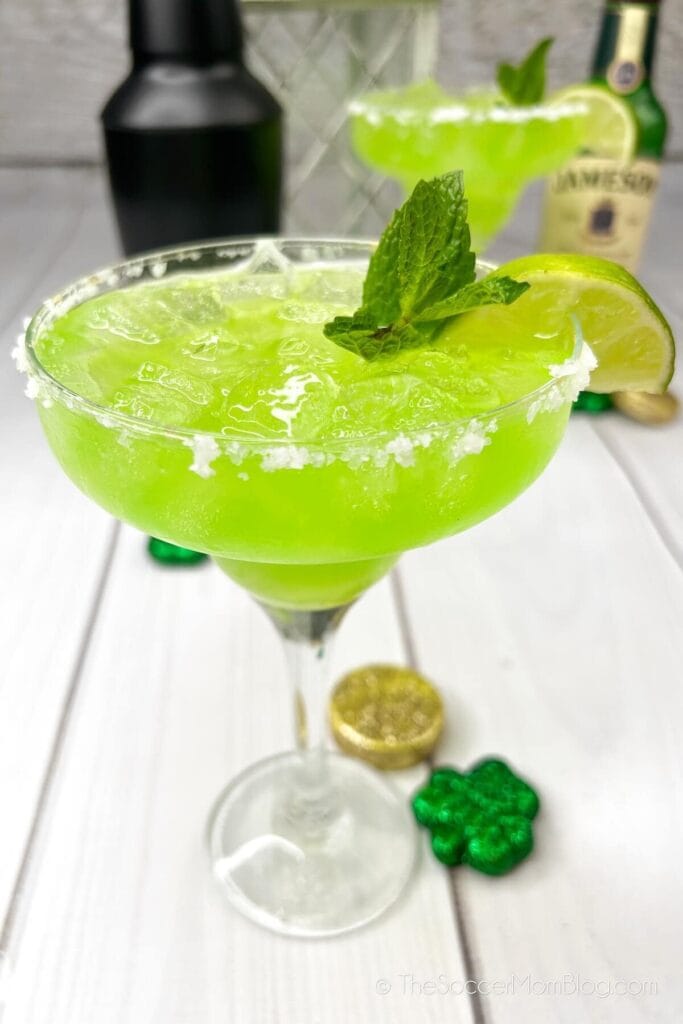 A margarita glass filled with a vibrant green drink, garnished with a lime wedge and fresh mint, rimmed with salt, and surrounded by festive decorations like shamrocks and gold coins on a white wooden table.