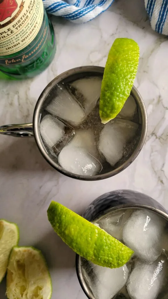 Overhead view of two metal mugs filled with a refreshing cocktail on ice, each garnished with a lime wedge, set on a marble surface with additional lime slices and a bottle of green liquor in the background.