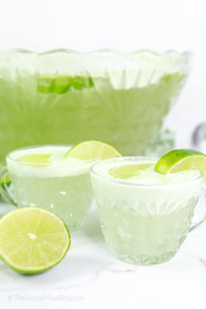 A large glass punch bowl filled with light green lime punch, accompanied by two matching cups garnished with lime slices, set on a white surface with a fresh lime cut in half for decoration.