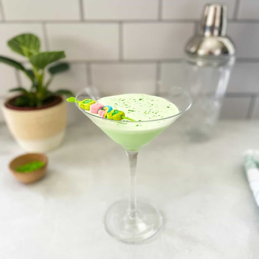 Martini glass filled with a creamy green drink, garnished with colorful marshmallows on a skewer and sprinkled with green decorations, set on a light-colored countertop with blurred kitchen accessories in the background.