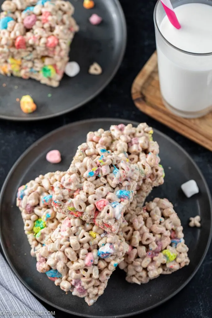 Lucky Charms and marshmallow cereal bars featuring colorful marshmallow bits, stacked on a black plate next to a glass of milk, perfect for festive treats or St. Patrick's Day celebrations.