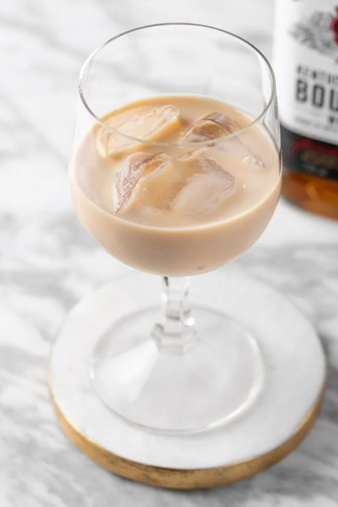 A stemmed glass filled with a creamy cocktail featuring ice cubes, offering a decadent boozy treat, placed on a white marble coaster with a blurred bourbon bottle in the background.